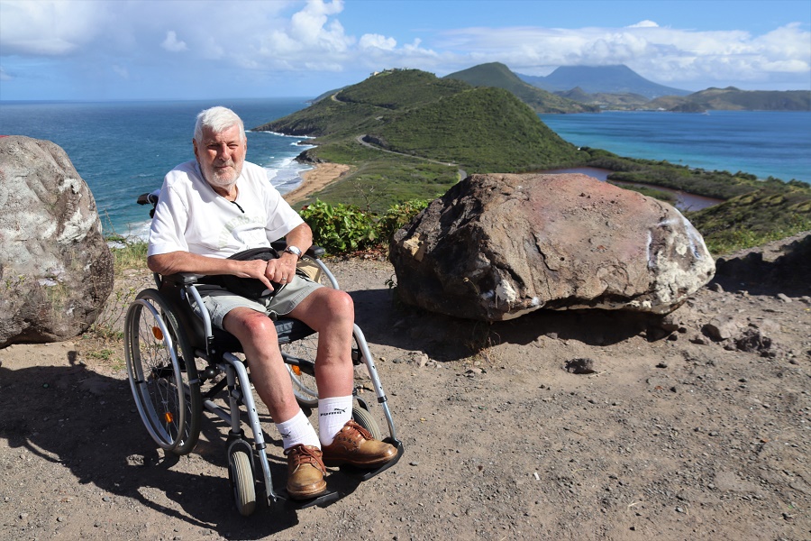 Jozef, zittend in een manuele rolstoel, poseert voor een panorama van groene heuvels, gouden stranden en een azuurblauwe oceaan.