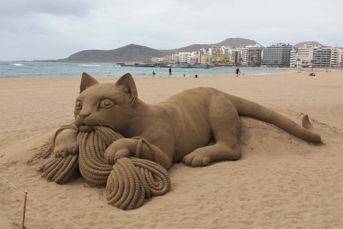 Zandsculptuur van een kat die met een tros touw speelt, op het strand.