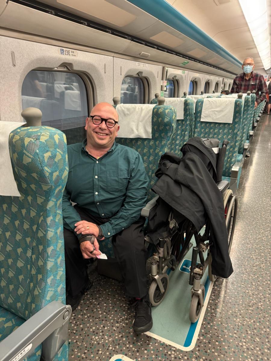 Interieur van de hogesnelheidstrein. Pieter heeft zijn manuele rolstoel dichthgeplooid en zit in de stoel ernaast.