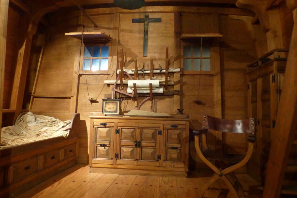 Een rek met papierrollen boven een houten kast, een leren stoel en een bed waar Christoffel Columbus net uitgestapt lijkt.