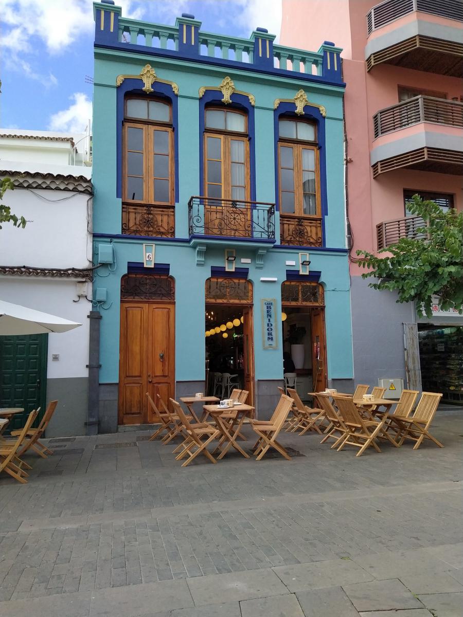 Achter deze blauwe historische gevel met hoge ramen schuilt Café Benidorm. Ervoor ligt een terras met houten meubels.
