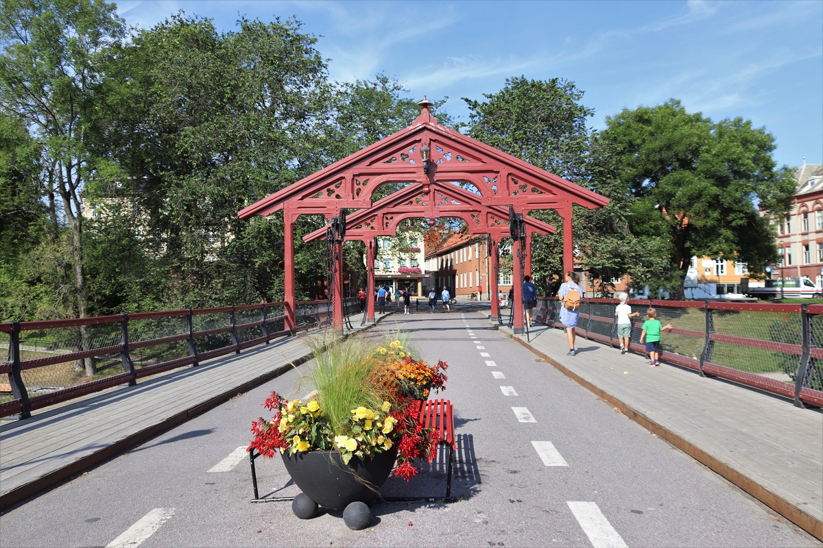 Wandelaars kuieren over de roodgeschilderde brug. Een bloembak verspert de weg voor autoverkeer.