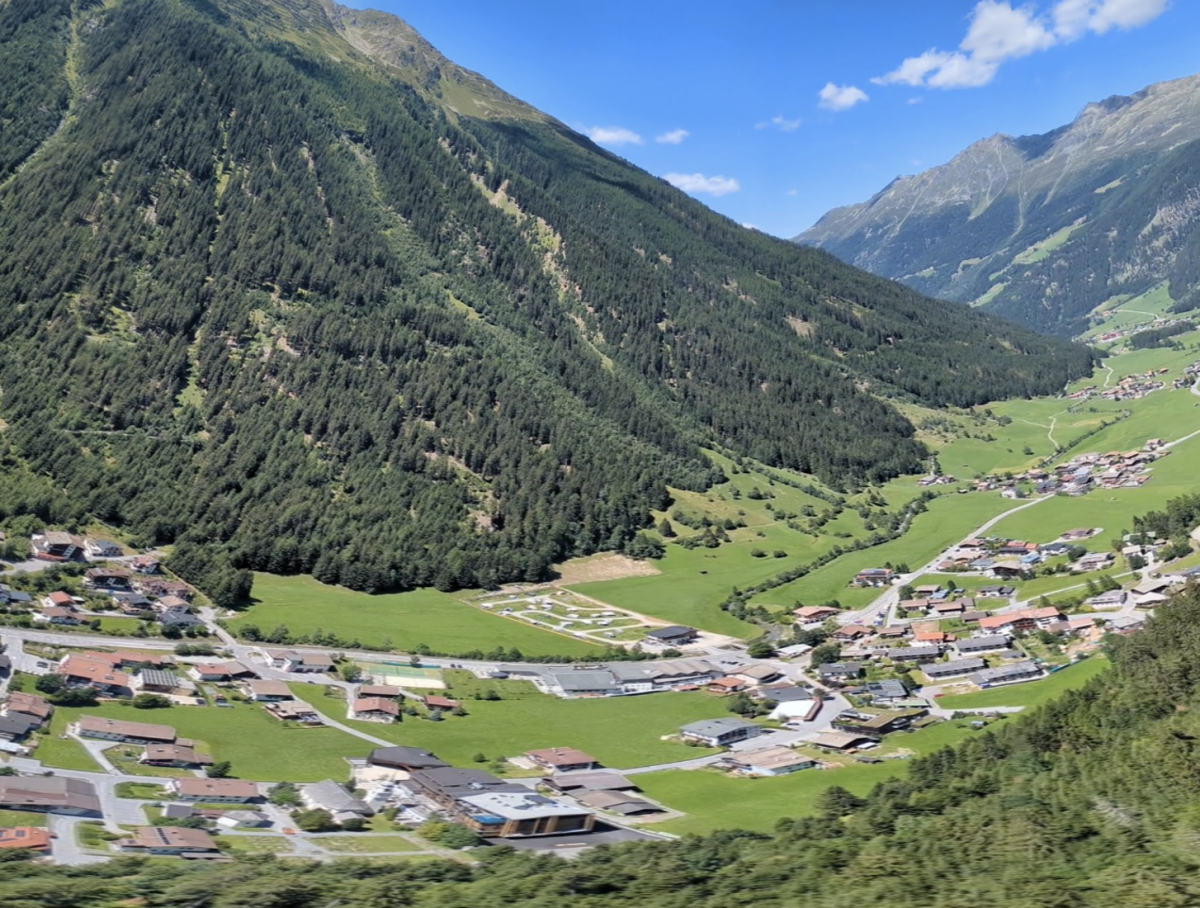 Panorama met gigantische beboste bergen en in vergelijking piepkleine huisjes in het (zomers) groene dal.