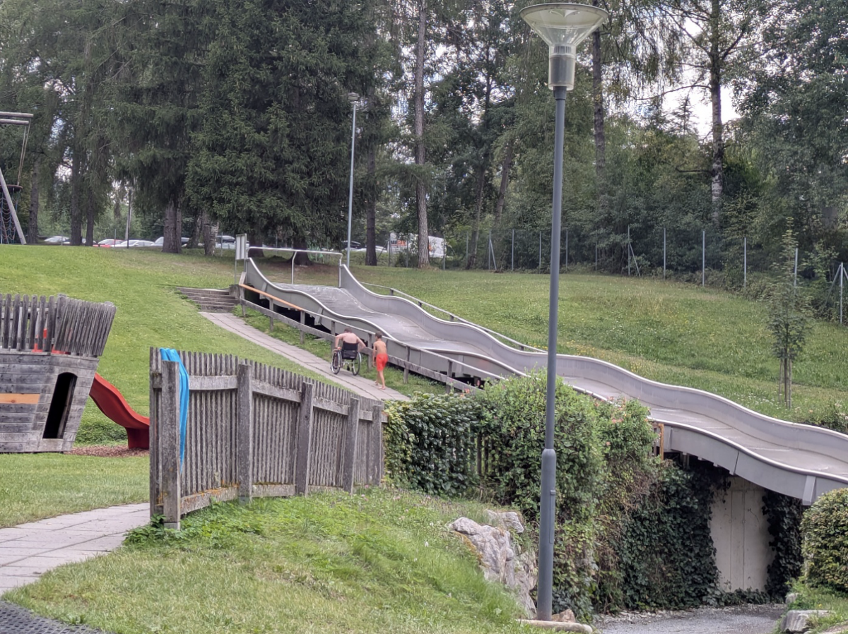 We zien van op afstand hoe vader (met rolstoel) en zoon samen een helling trotseren om naar de glijbaan te gaan.