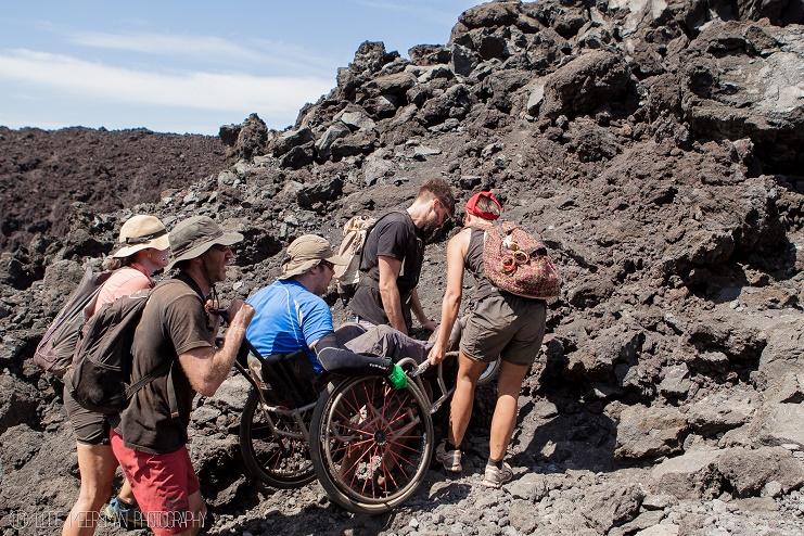 Een rolstoeler klimt met hulp van vier reisgenoten een rots op.