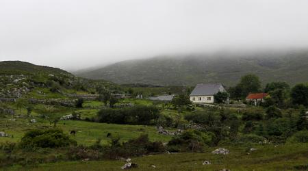 Onder een mistig grijze hemel zijn een paar huisjes tussen de groene heuvels gestrooid.